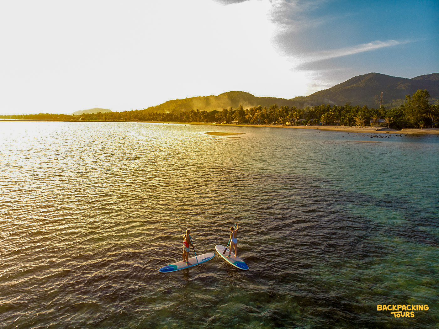 Two people paddleboard across the glistening ocean at sunset near the shores of Koh Phangan.