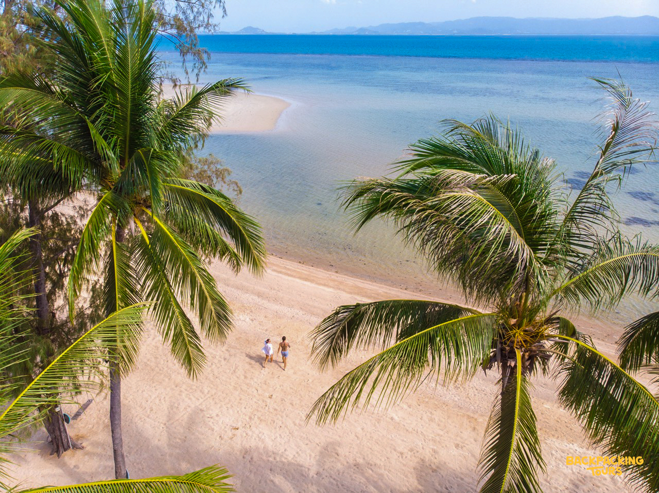 Two people stroll along a beautiful sandy beach with clear ocean and palm trees in Koh Phangan.