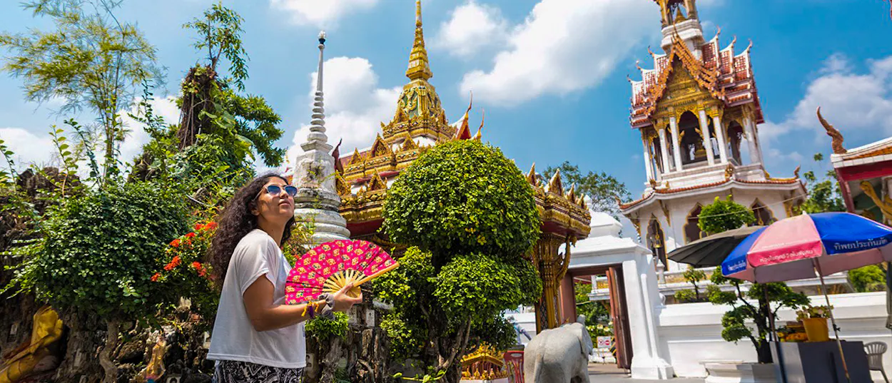 A woman holding a fan looks up at the ornate golden temples and white stupas in Bangkok.