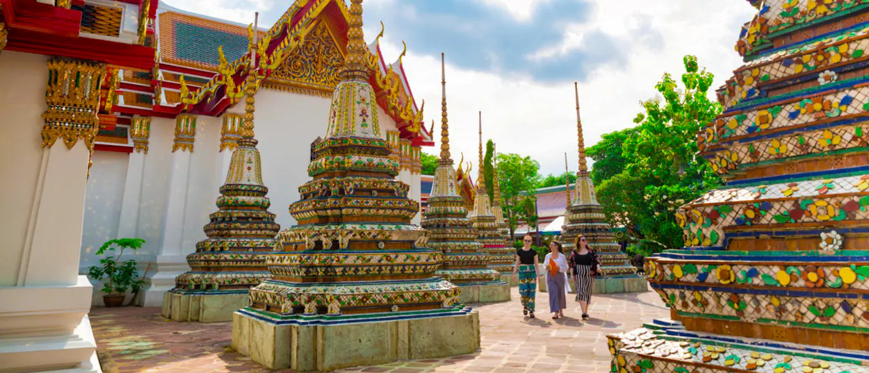 Three women explore the ornate golden chedis and temple grounds of Wat Pho in Bangkok.
