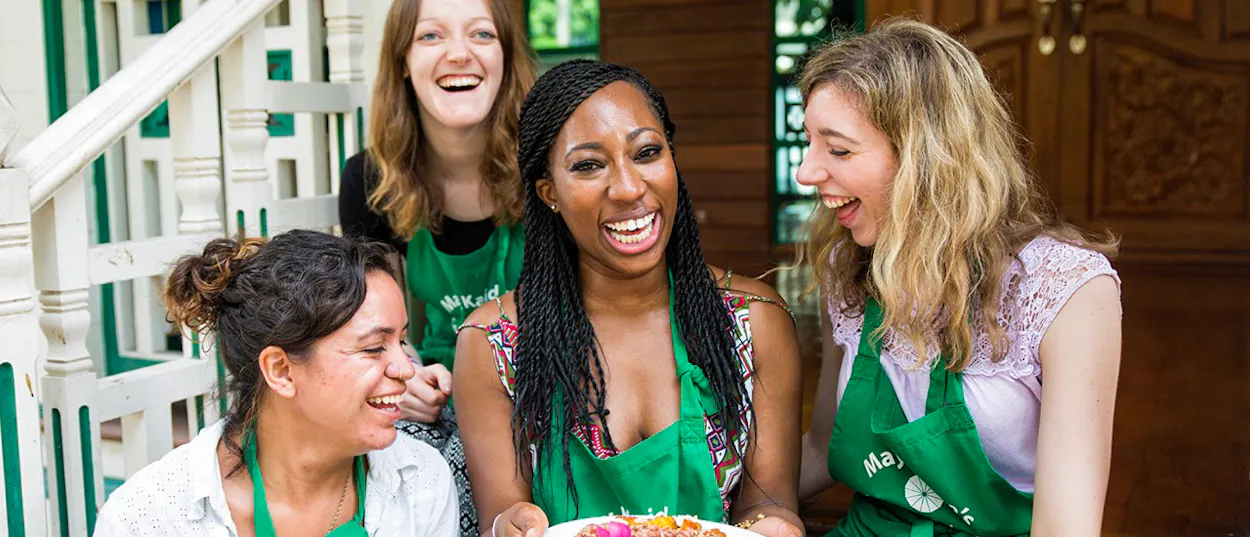 Four happy women in green aprons laugh while holding a plate of food at a Bangkok cooking class.
