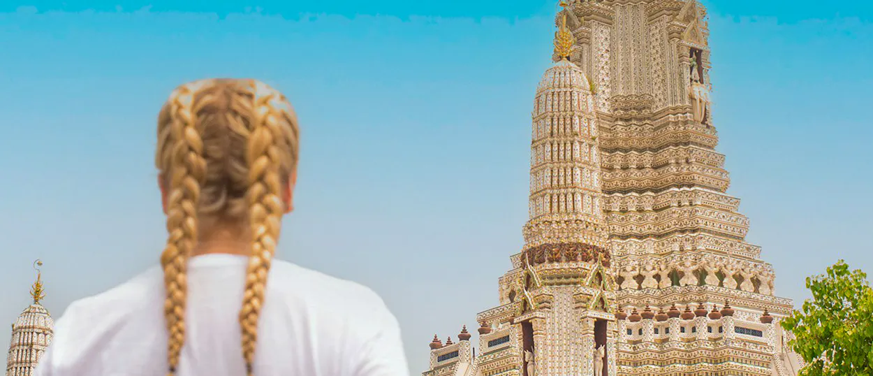 Blonde-braided traveler views the majestic, intricately carved spires of Wat Arun temple in Bangkok.