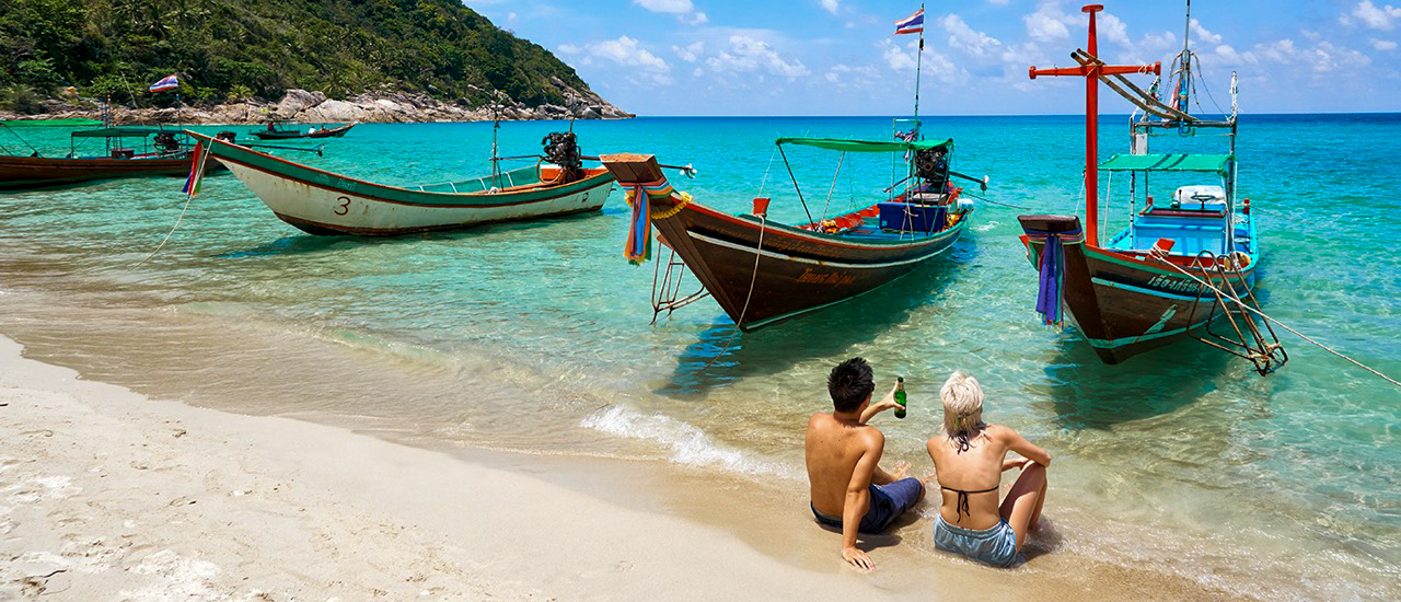 Two people sit on a sandy beach, watching longtail boats in clear turquoise water at Koh Phangan.