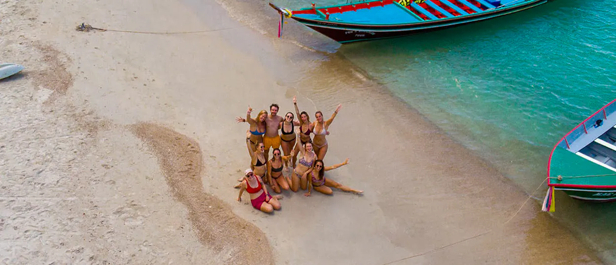 A joyful group poses on Koh Phangan beach with turquoise water and traditional longtail boats.