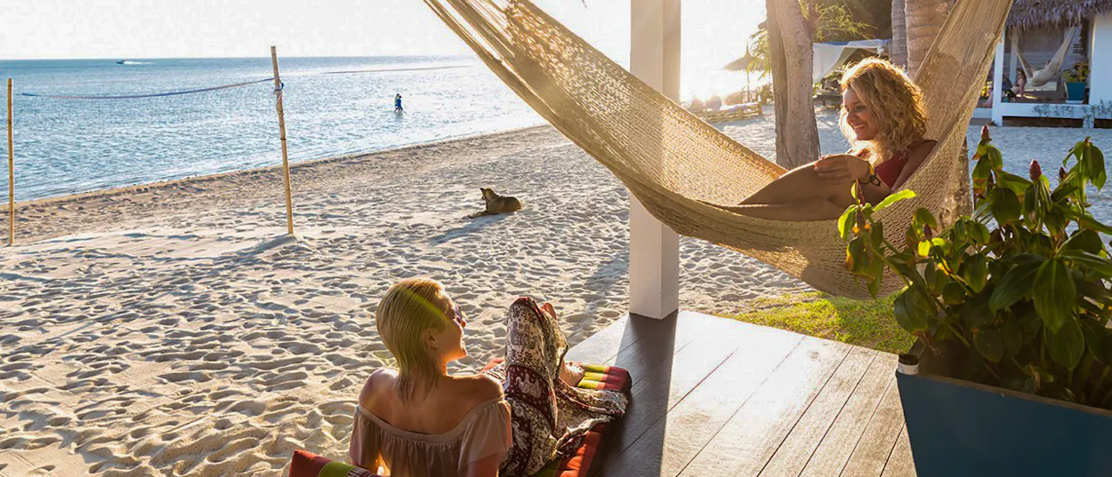 Two women relax on a beachside porch, one in a hammock, enjoying the sunny Koh Phangan beach.