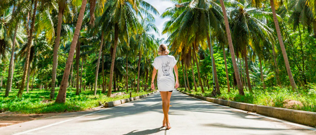 A woman walks down a sunny road lined with tall palm trees on Koh Phangan.