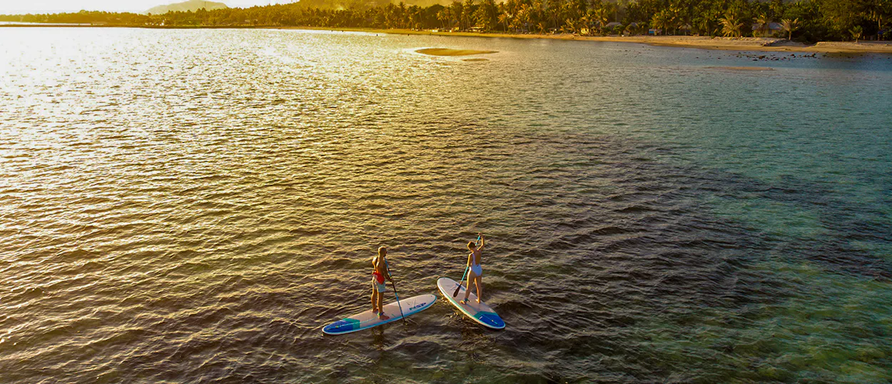 Two people paddleboard on calm, golden water near the lush coast of Koh Phangan at sunset.