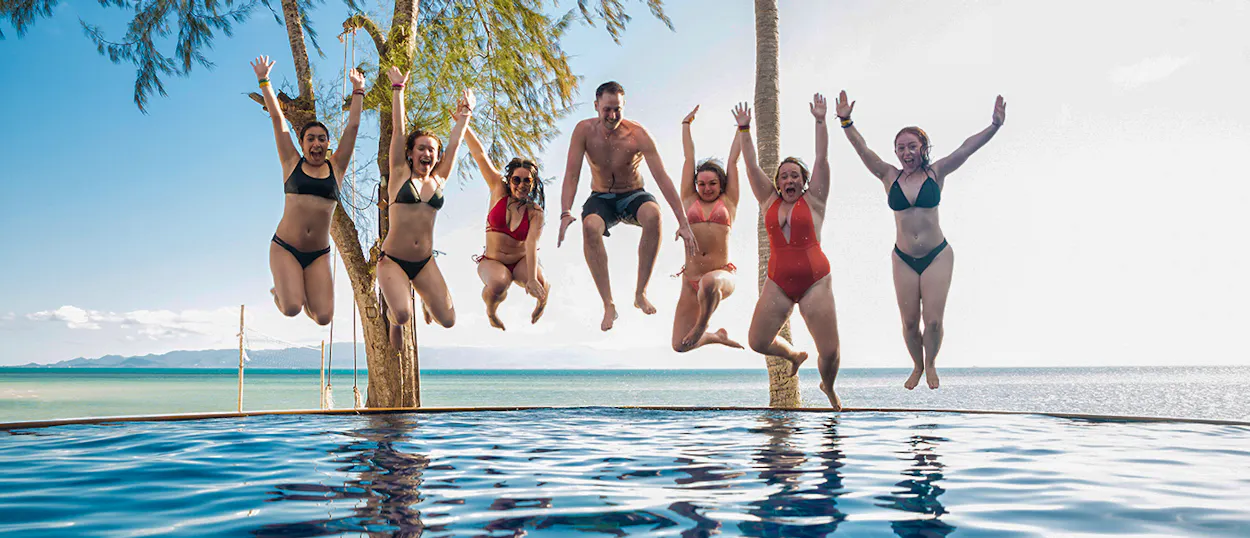 Seven happy friends jump together above an infinity pool overlooking the sea in Koh Phangan.