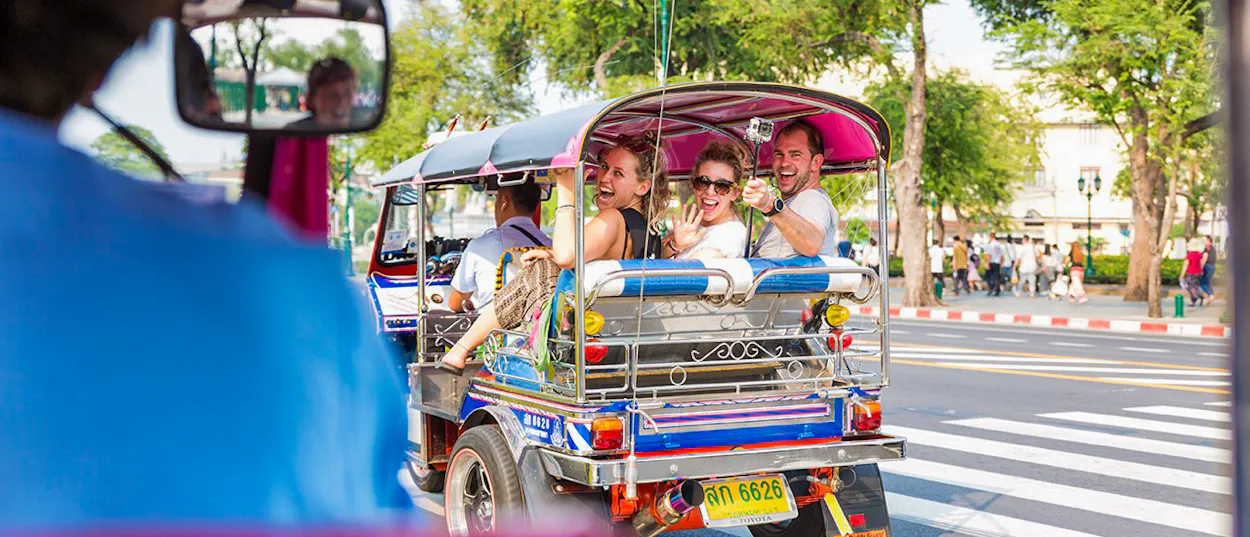 Tourists smiling and waving from a traditional tuk-tuk on a sunny Bangkok street.
