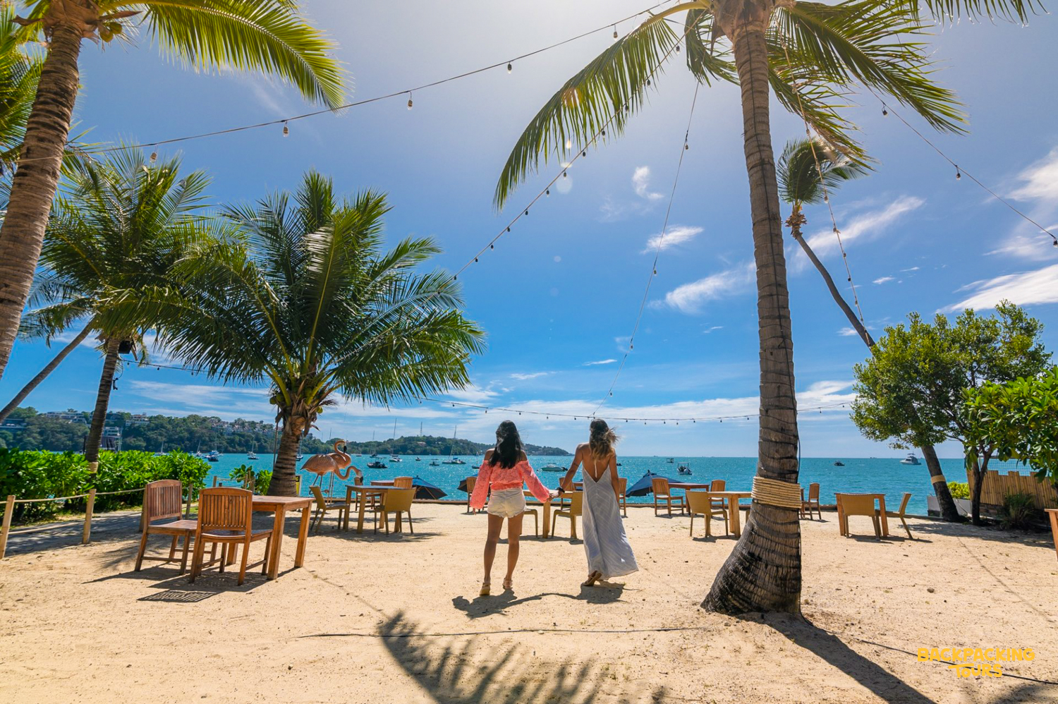 Two women stroll hand-in-hand on a sunny Phuket beach towards the beautiful blue ocean.