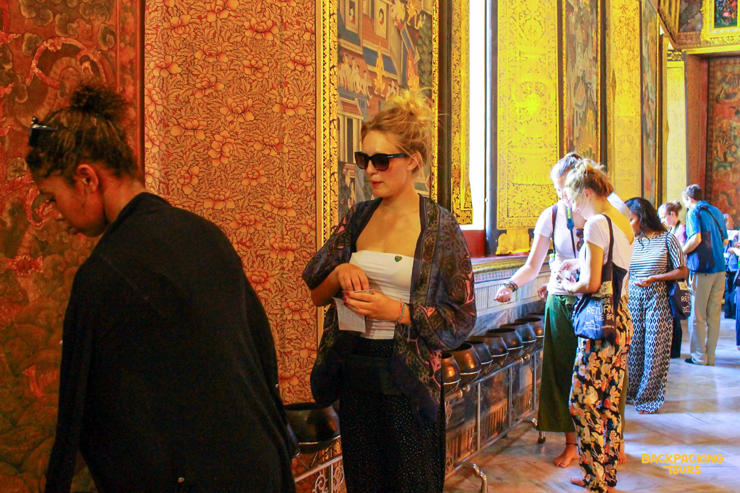 Travelers observing the ornate wall paintings and architecture within a Thai temple in Bangkok.