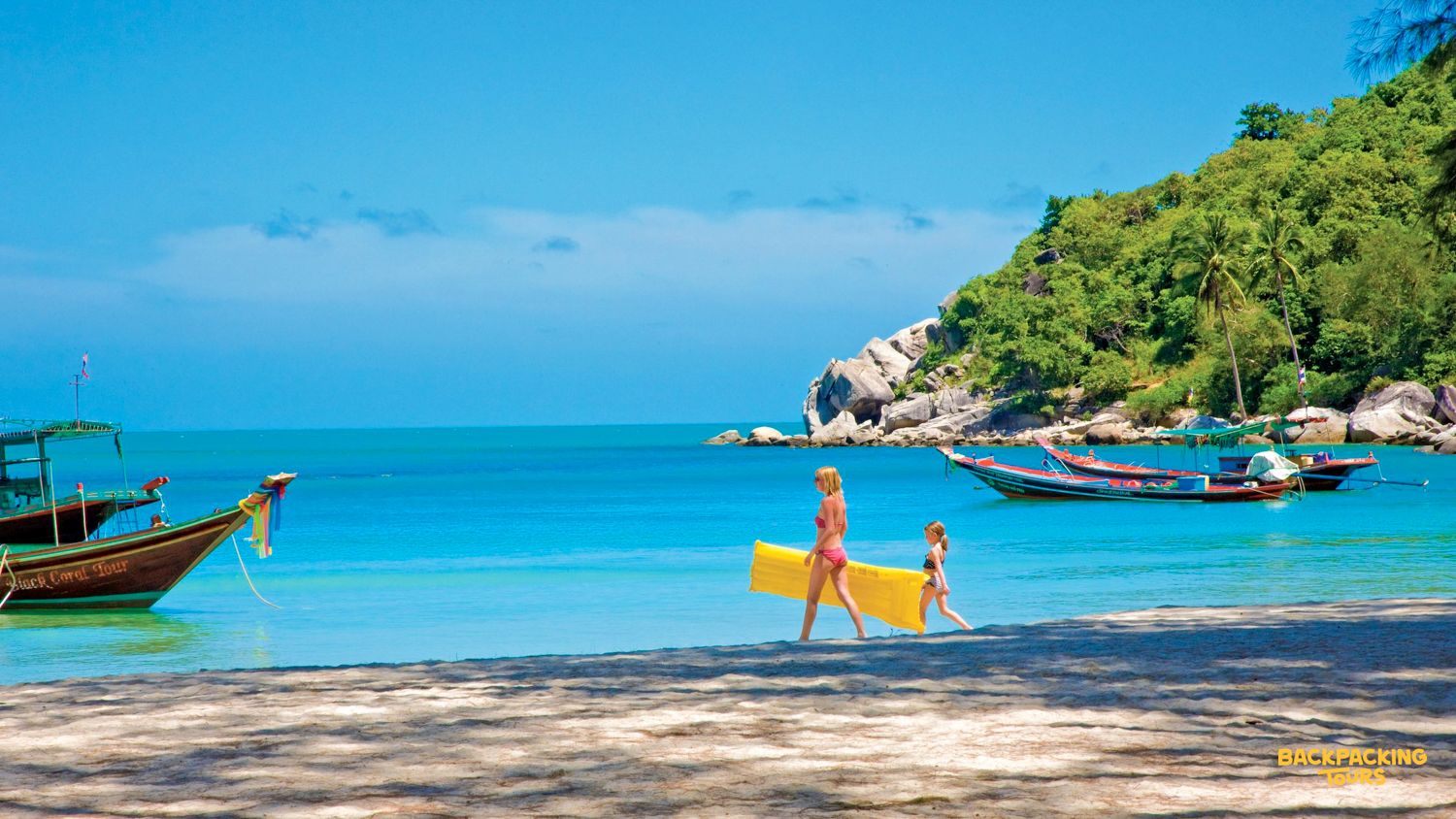 A woman and child walk along a sunny beach, carrying a yellow raft on Koh Phangan.