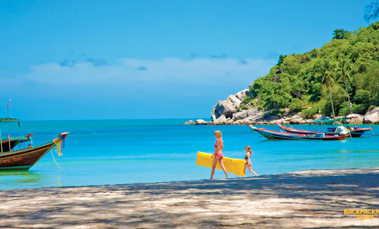 A woman and child walk along a sunny beach, carrying a yellow raft on Koh Phangan.