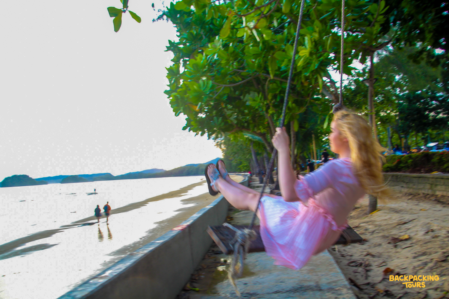 A blonde woman in a pink dress joyfully swings under trees on a beautiful beach in Krabi.