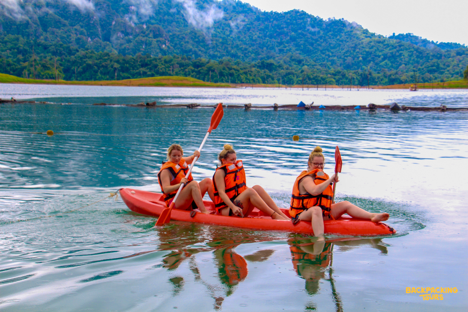 Three friends paddle a red kayak across the serene lake in Thailand's Khao Sok National Park.