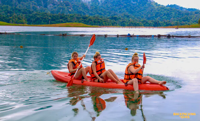 Three friends paddle a red kayak across the serene lake in Thailand's Khao Sok National Park.