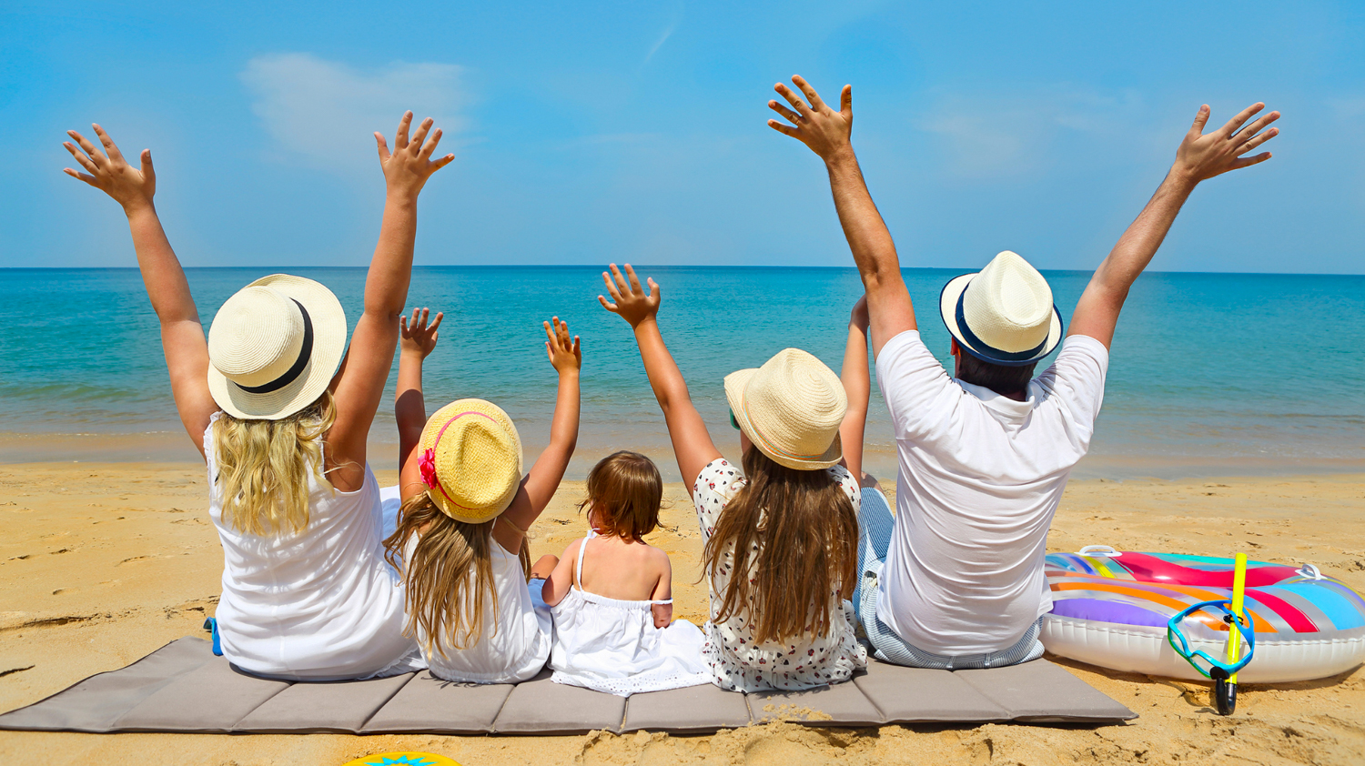 A joyful family sits on a sandy beach, raising hands to the clear blue ocean in Thailand.