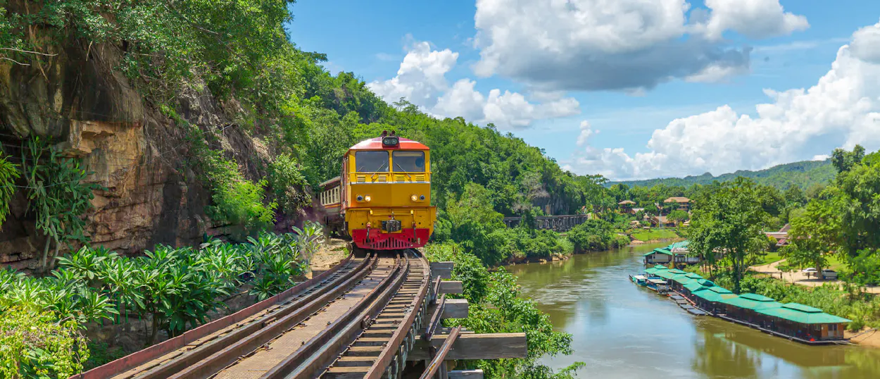 A yellow and red train travels along a scenic track beside a tropical river in Thailand.