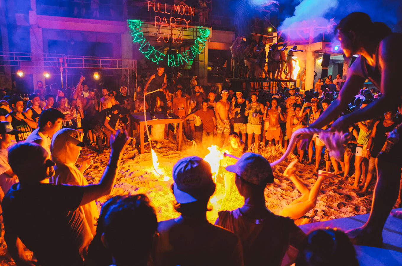 Revellers dance around a large fire pit at a vibrant Full Moon Party on a beach at night.