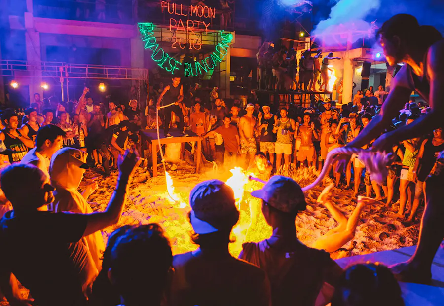Revellers dance around a large fire pit at a vibrant Full Moon Party on a beach at night.