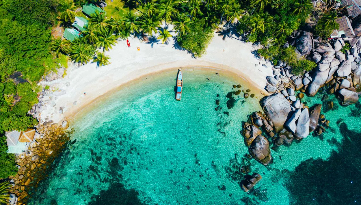 Tropical white sand beach with a boat, clear turquoise water, and palm trees on Koh Phangan.