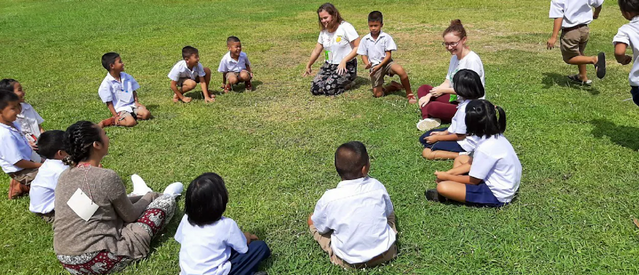Two volunteers teach a group of young Thai children sitting in a circle on green grass.