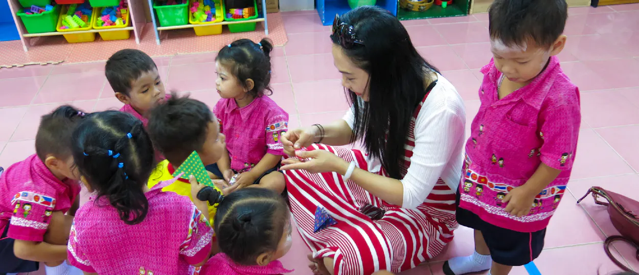 A volunteer teacher sits on the floor, engaging with young children in a Thai kindergarten.