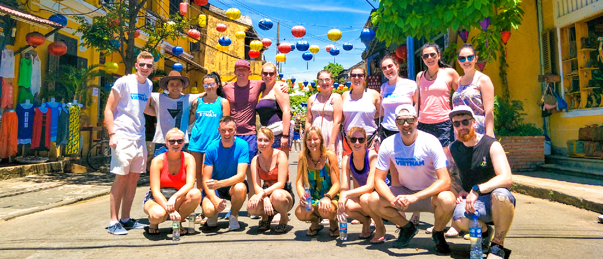Happy backpackers pose on a sunny street in Hoi An, Vietnam, with colorful lanterns overhead.