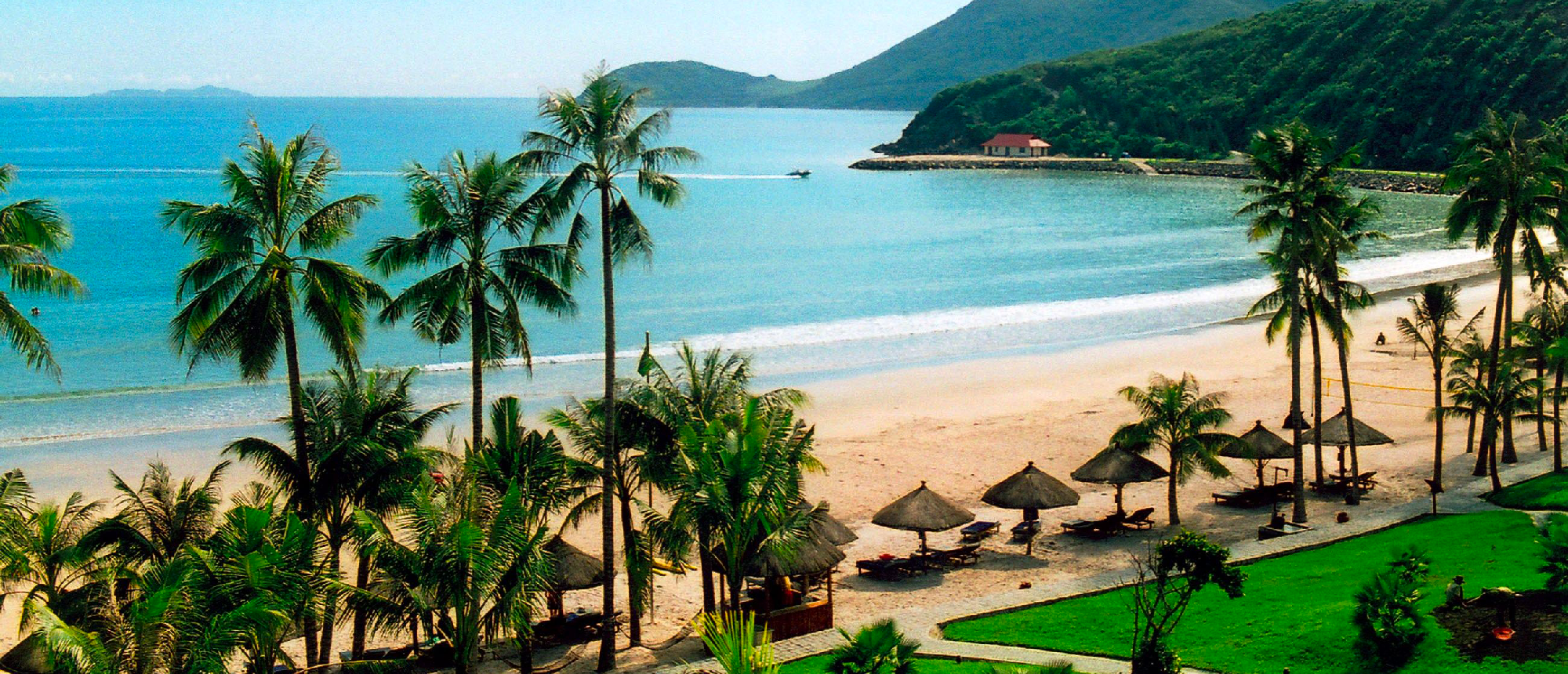 Beautiful Mui Ne beach with palm trees, straw umbrellas, and calm turquoise water, backed by green mountains.