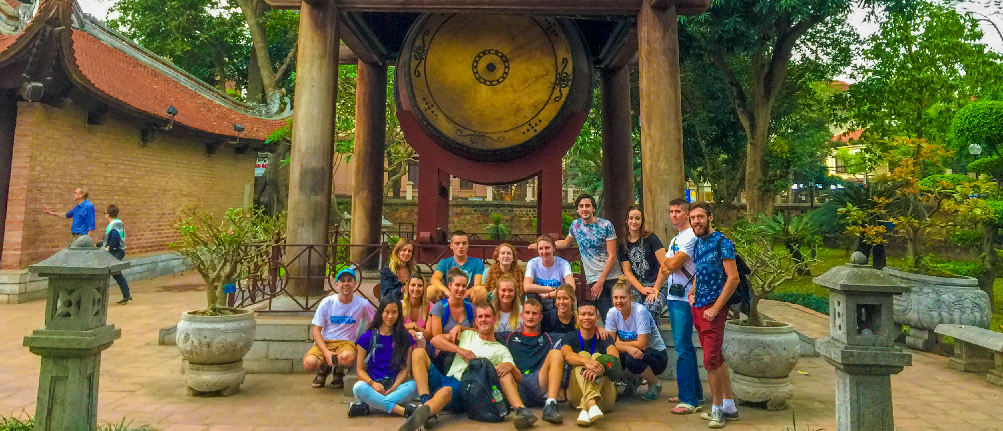 A large group of happy backpackers poses in front of a giant traditional drum at a temple in Hanoi.