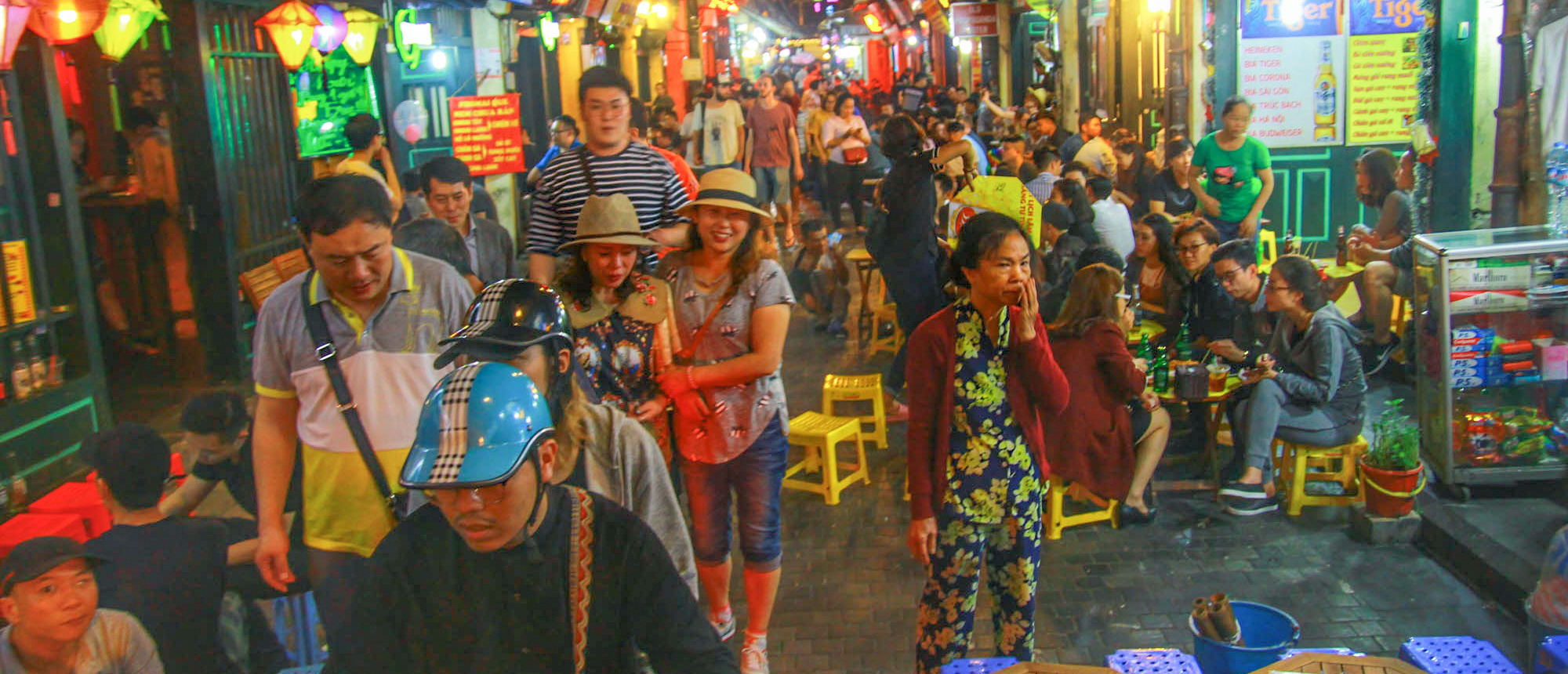 A bustling night market street in Hanoi, illuminated by colorful lanterns, with many people walking and dining.