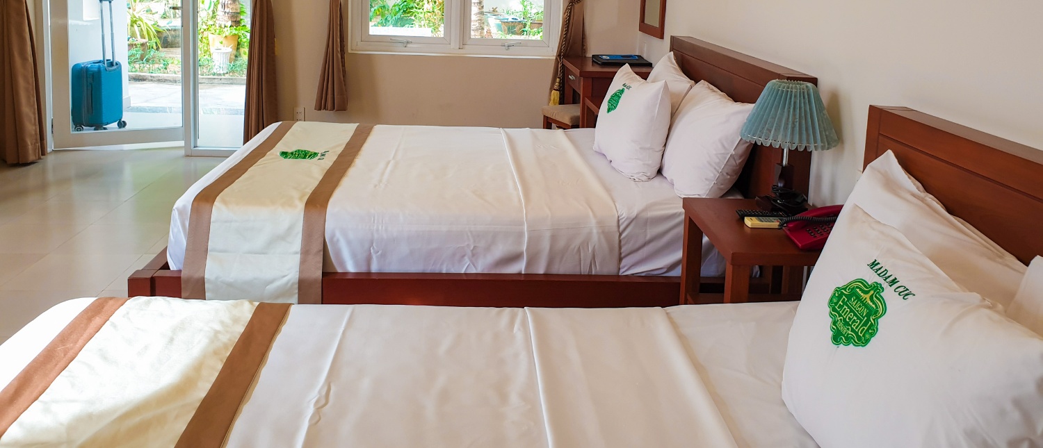 A bright hotel room in Mui Ne with two beds, a lamp, and a blue suitcase.