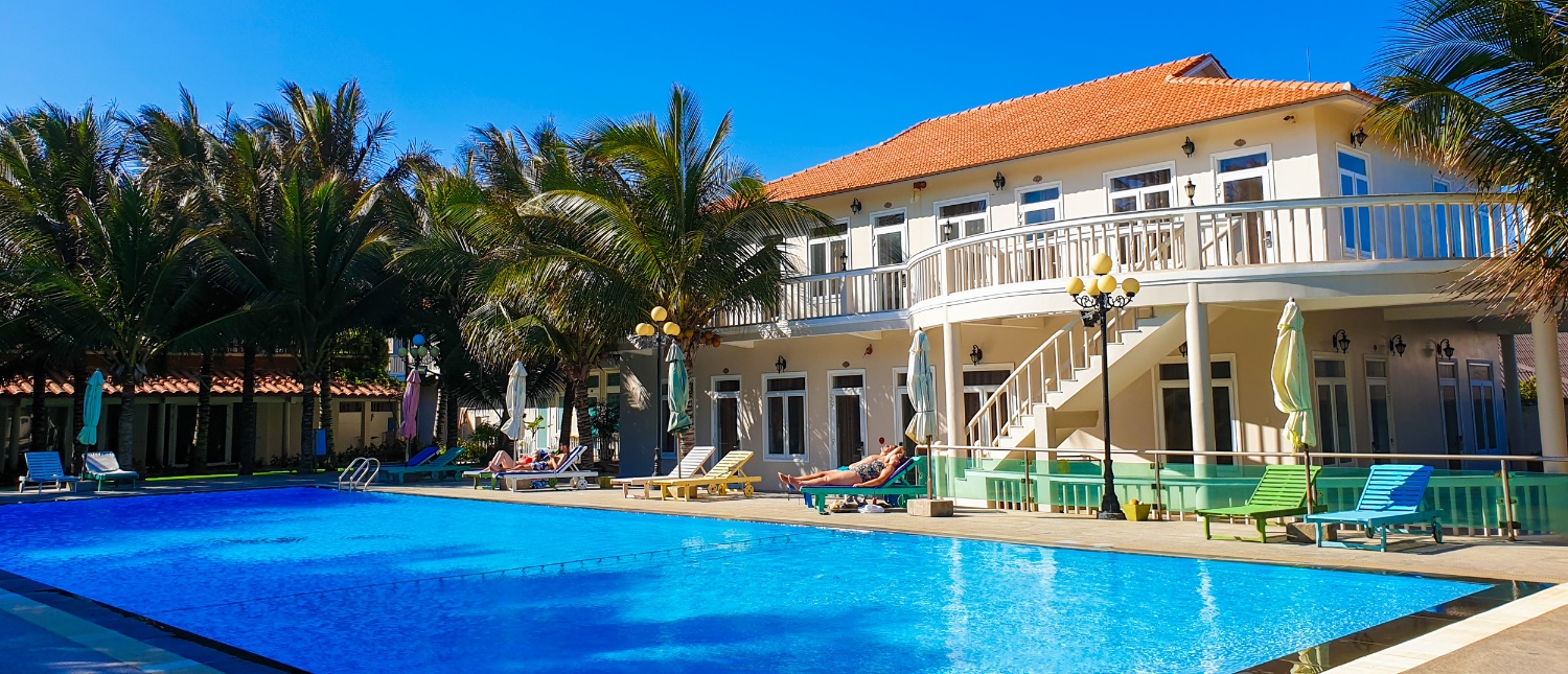 Guests relax by a large blue swimming pool at a sunny Mui Ne resort with palm trees.