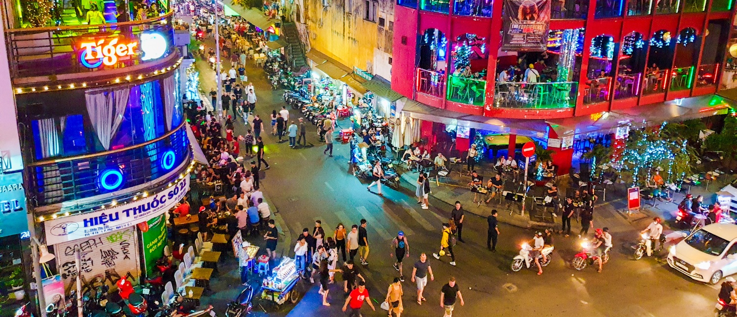 Lively street scene at night in Ho Chi Minh with many people, motorbikes, and bright neon lights.