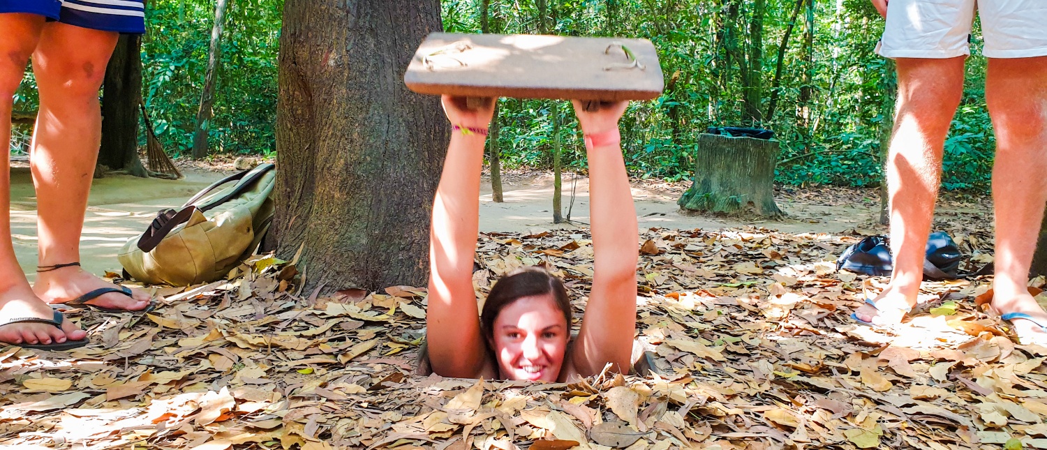 Smiling woman emerging from a camouflaged tunnel entrance in a leafy forest, with other tourists watching.