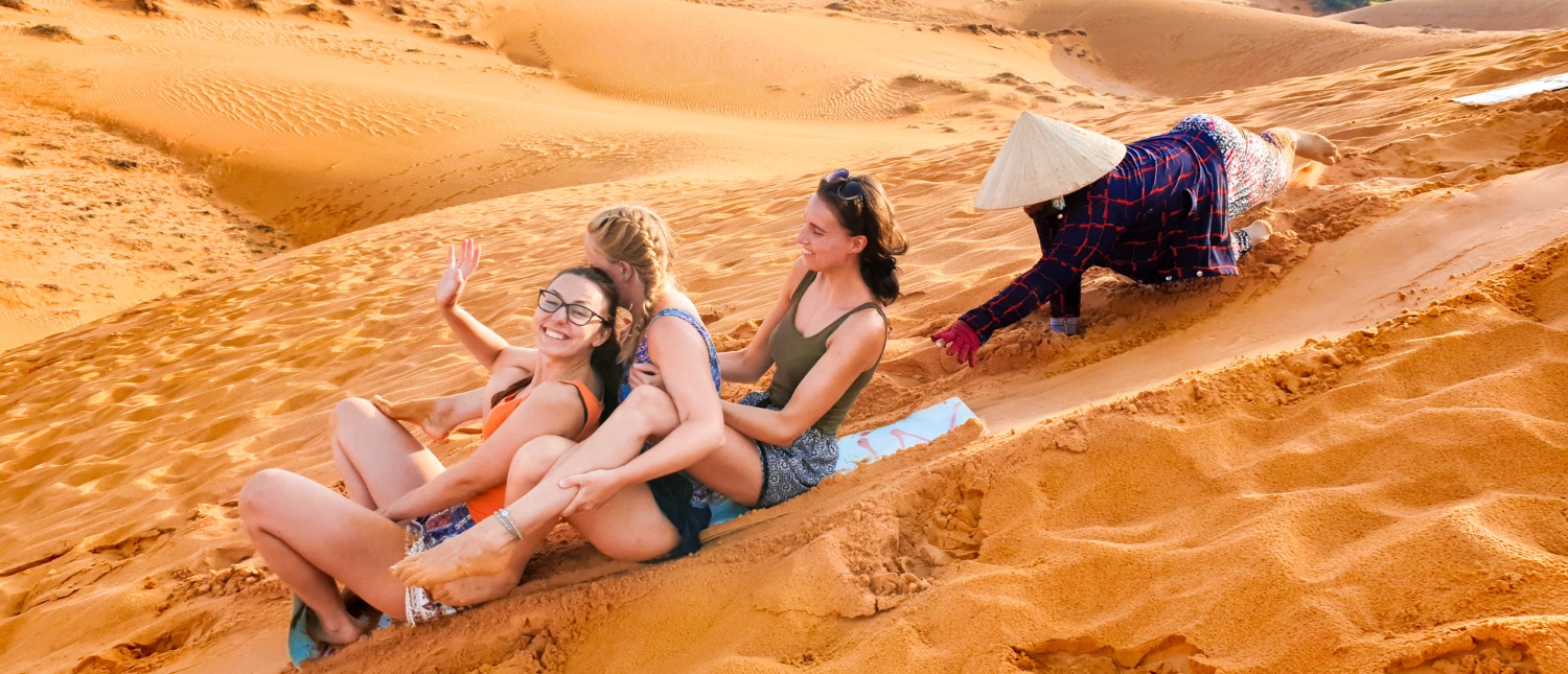 Three happy women sand sledding down golden dunes in Mui Ne, Vietnam, pushed by a local.