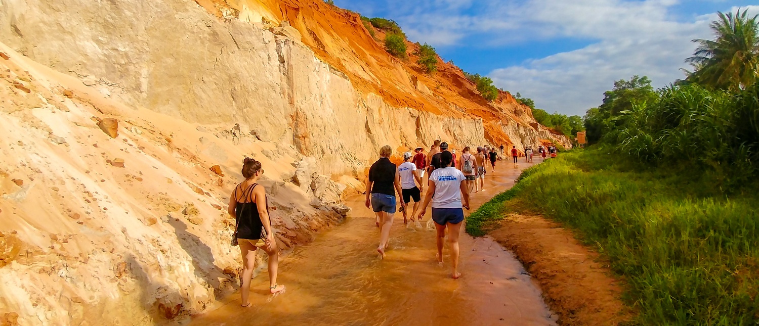 Travelers explore the enchanting Fairy Stream in Mui Ne, walking barefoot between colorful eroded banks and green vegetation.