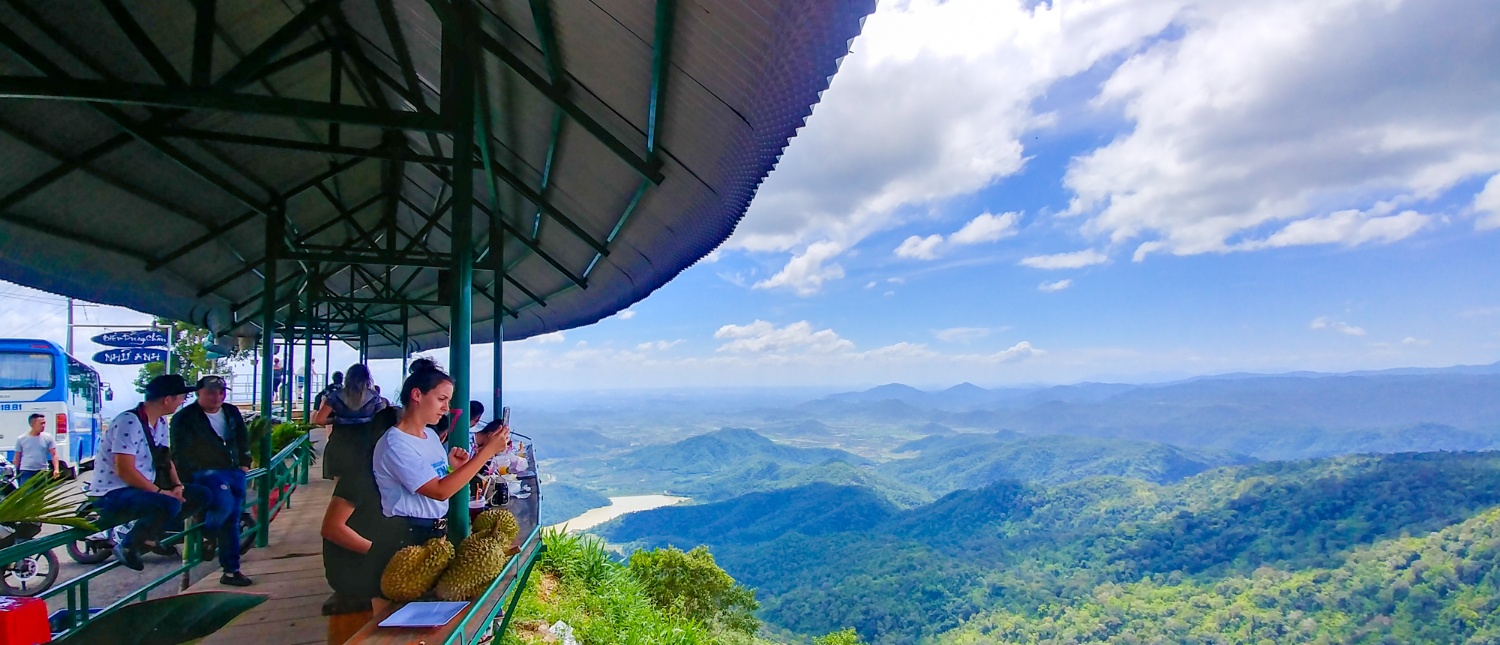Travelers at a viewpoint overlooking lush green mountains, a river, and a blue sky in Vietnam.