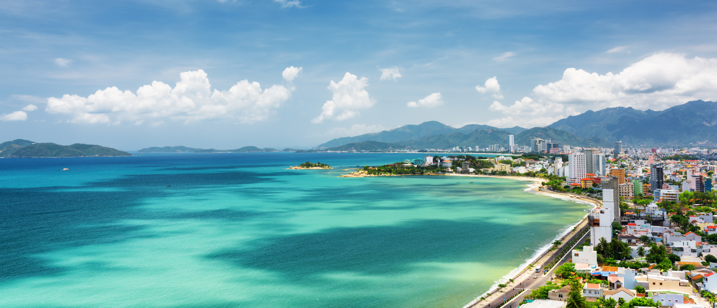 Nha Trang's vibrant coastline features clear turquoise water, a sandy beach, and city buildings against mountains.