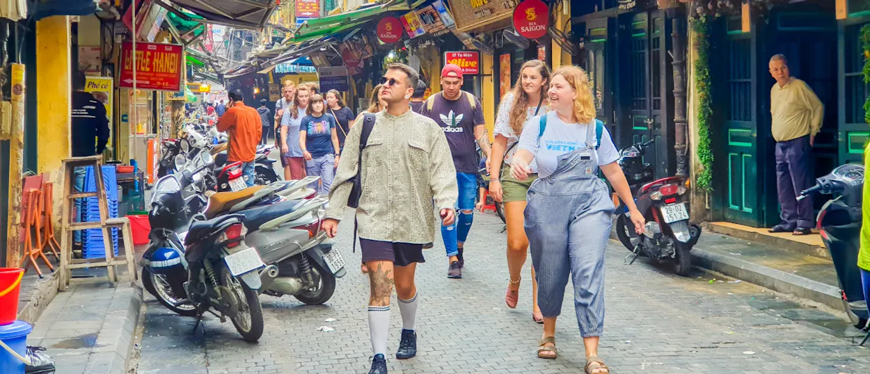 Tourists walk through a busy, narrow street in Hanoi, past parked motorbikes and colorful shops.