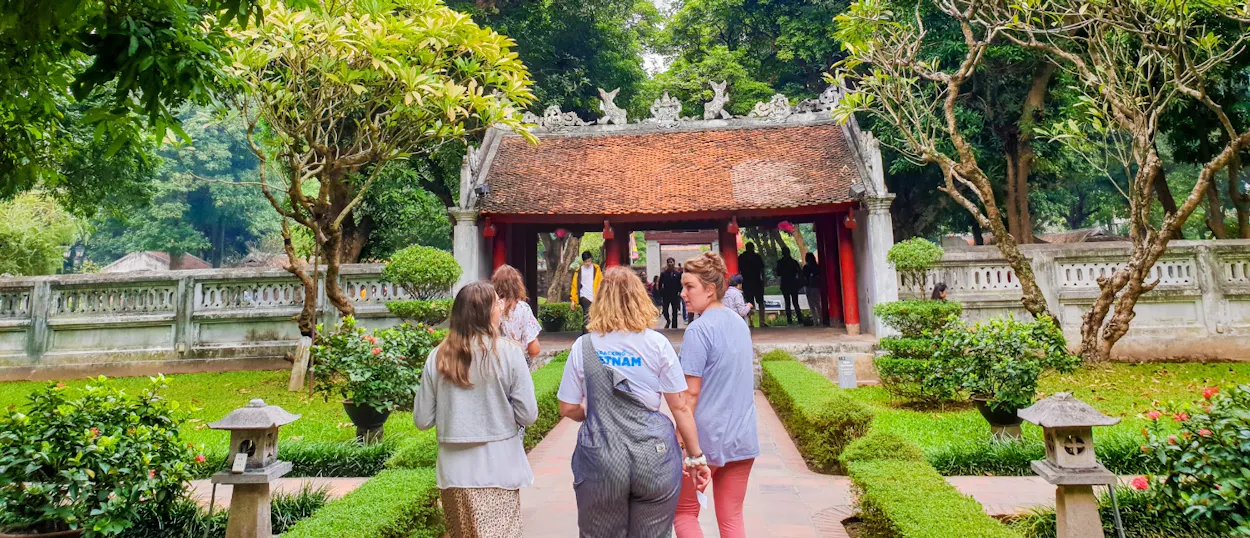 Four women walk through a lush garden towards a traditional temple gate in Hanoi.
