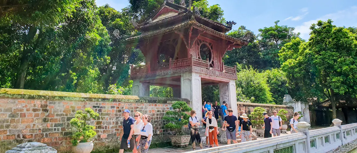 Tourists walk past an ancient red wooden pavilion and brick wall surrounded by green trees in Hanoi.