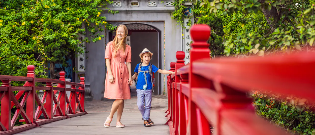 A smiling woman and a boy walk hand-in-hand on the vibrant red Huc Bridge in Hanoi.