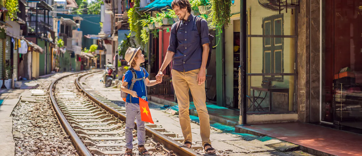 A smiling father and child hold hands, walking along train tracks in a lively Hanoi street.