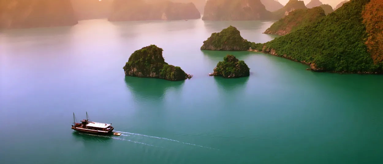 A traditional boat cruises through the emerald waters of Halong Bay at sunset, surrounded by limestone islands.