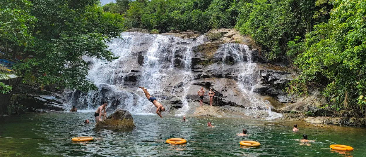 People swim and dive in a refreshing multi-tiered waterfall pool surrounded by lush green trees in Vietnam.
