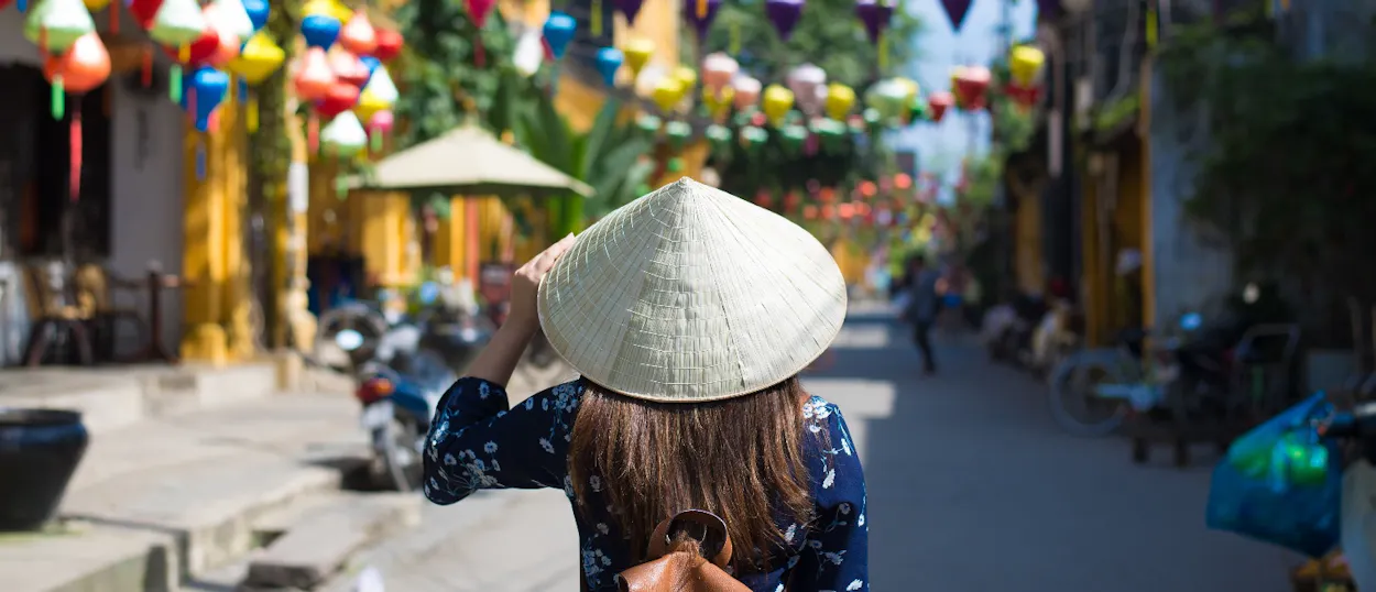 A person in a conical hat walks through a lively street in Hoi An adorned with colorful lanterns.