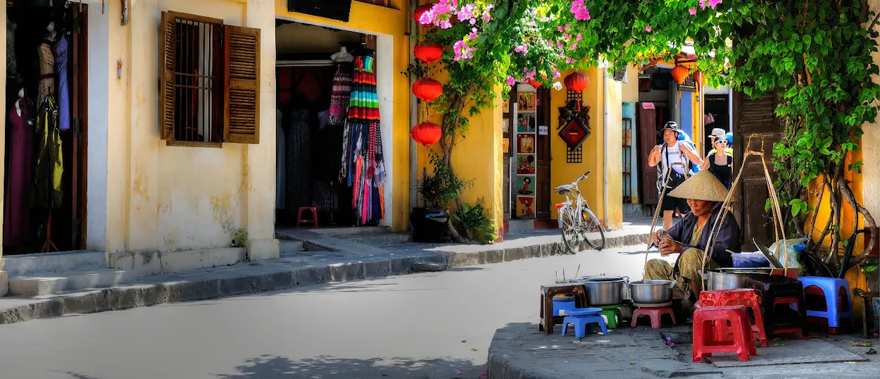 A person in a conical hat sells street food under flowering vines and red lanterns in Hoi An.