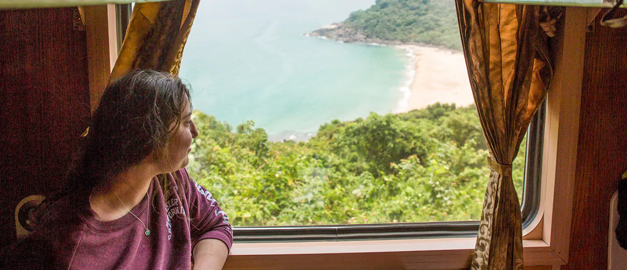 Woman on a train looks out at a beautiful beach, green hills, and turquoise ocean.