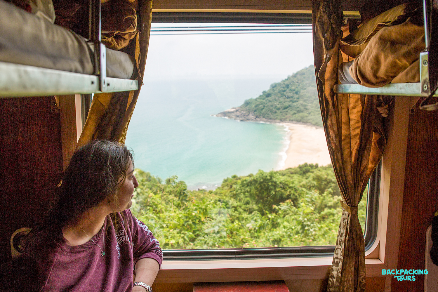 A woman gazes from an overnight train window at a beautiful Vietnamese coastline with a sandy beach.